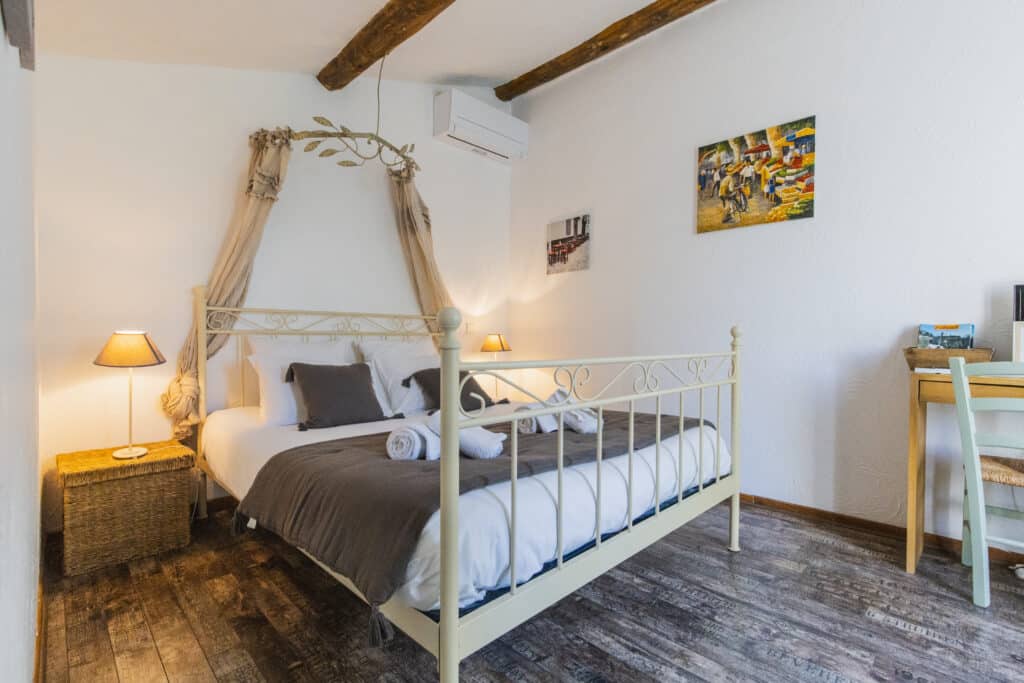 Chambre sous les combles avec lit en fer forgé et baldaquin, poutres apparentes et murs blancs
