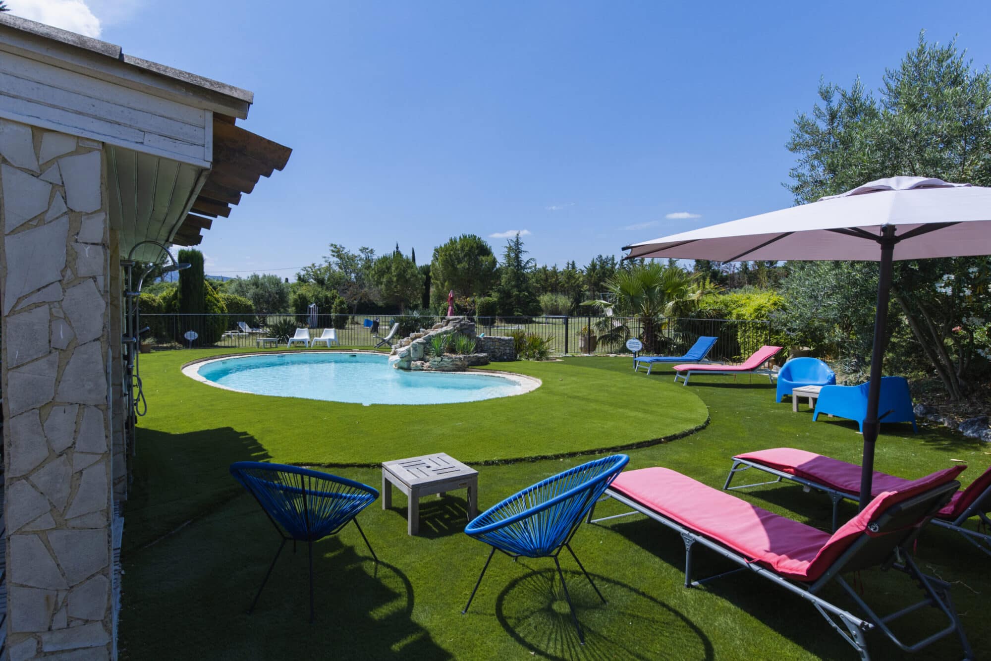 Piscine ovale entourée d’un jardin avec pelouse, transats roses et fauteuils bleus, sous un ciel dégagé, en Luberon
